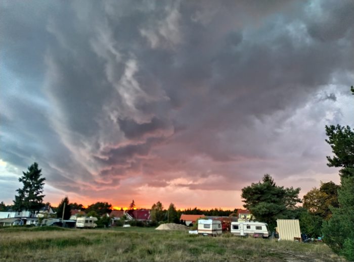ein Sonnenuntergang hinter einer Dorfkulisse, darüber mäandern dunkle Gewitterwolken, denkt an Caspar David Friedrich